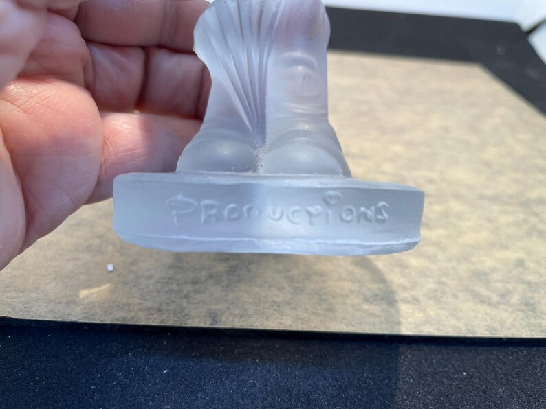 May include: A clear glass figurine of a person wearing a hat and boots. The figurine is standing on a base that says "Productions".