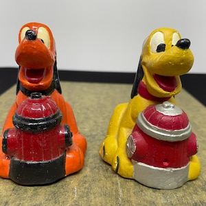 May include: Two ceramic figurines of Pluto the dog from Disney, holding red fire hydrants. One Pluto is orange and the other is yellow.