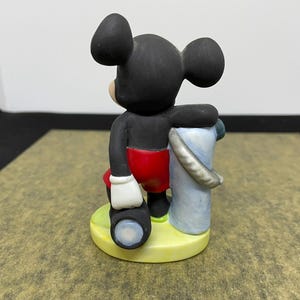 May include: A ceramic figurine of Mickey Mouse, wearing red shorts, white gloves, and black shoes. Mickey is holding a telescope and a blue and white bucket. The figurine is on a yellow base.