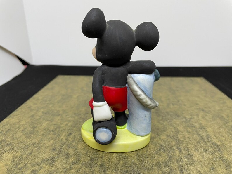 May include: A ceramic figurine of Mickey Mouse, wearing red shorts, white gloves, and black shoes. Mickey is holding a telescope and a blue and white bucket. The figurine is on a yellow base.