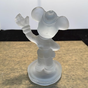 May include: A frosted glass figurine of Mickey Mouse wearing a hat and holding his glove up in the air. The figurine is standing on a round base.