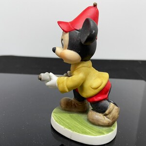 Puede incluir: Una figurita de cer&aacute;mica de Mickey Mouse con un sombrero rojo y una chaqueta amarilla. Est&aacute; sosteniendo un palo de golf y de pie sobre una base verde.
