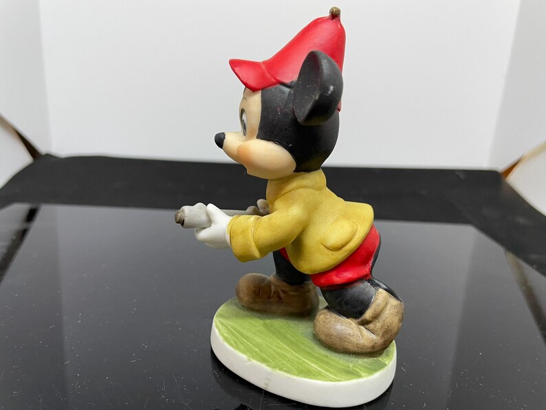Puede incluir: Una figurita de cer&aacute;mica de Mickey Mouse con un sombrero rojo y una chaqueta amarilla. Est&aacute; sosteniendo un palo de golf y de pie sobre una base verde.