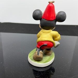Puede incluir: Una figurita de cer&aacute;mica de Mickey Mouse con un sombrero rojo y una camisa amarilla. Est&aacute; de pie sobre una base verde.