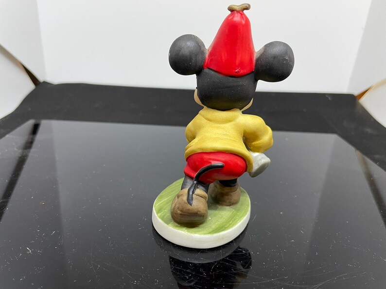 Puede incluir: Una figurita de cer&aacute;mica de Mickey Mouse con un sombrero rojo y una camisa amarilla. Est&aacute; de pie sobre una base verde.