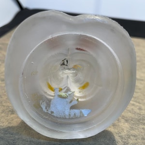 Puede incluir: Un tap&oacute;n de botella de vidrio transparente con un acabado esmerilado. El tap&oacute;n presenta un dise&ntilde;o floral blanco y amarillo con un peque&ntilde;o animal blanco en su interior.