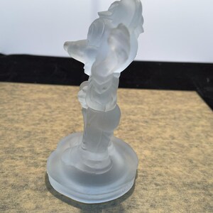 May include: A frosted glass figurine of a cartoon character standing on a round base. The character is wearing a hat and has a long nose.