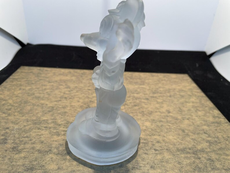 May include: A frosted glass figurine of a cartoon character standing on a round base. The character is wearing a hat and has a long nose.