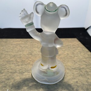 Puede incluir: Una figurita de vidrio esmerilado de Mickey Mouse con un sombrero y haciendo un saludo. La figurita est&aacute; de pie sobre una base de vidrio transparente.