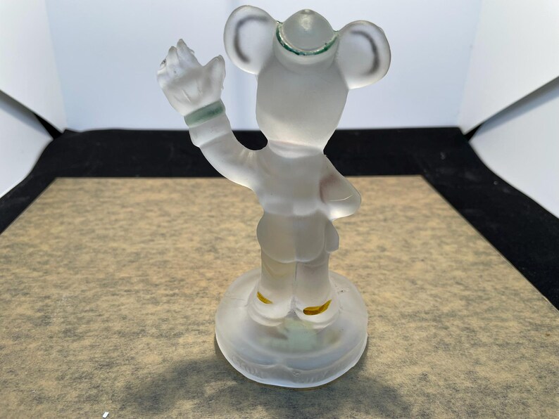 Puede incluir: Una figurita de vidrio esmerilado de Mickey Mouse con un sombrero y haciendo un saludo. La figurita est&aacute; de pie sobre una base de vidrio transparente.