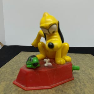 Könnte beinhalten: Eine gelbe und schwarze Comic-Hundefigur, die auf einem roten Sockel mit einer grünen Schildkrötenfigur sitzt. Der Hund schaut auf die Schildkröte. Der Sockel hat ein knochenförmiges Design.