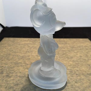 May include: A frosted glass figurine of Mickey Mouse standing on a circular base. The figurine is white and opaque.
