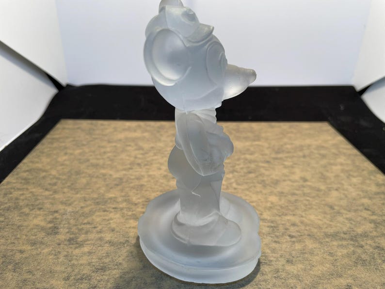 May include: A frosted glass figurine of Mickey Mouse standing on a circular base. The figurine is white and opaque.