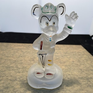 Puede incluir: Una figurita de cristal transparente de Mickey Mouse con un sombrero y un paraguas. La figurita est&aacute; pintada con detalles amarillos, rojos y azules.