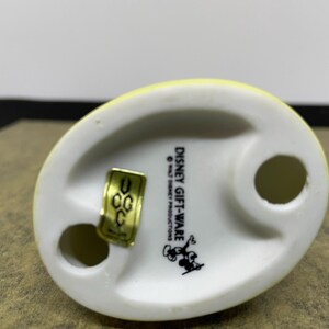 May include: A Disney Gift-Ware figurine base, featuring a yellow top and a white base with two holes. The base has the Disney Gift-Ware logo and a Mickey Mouse graphic. A gold sticker is attached.
