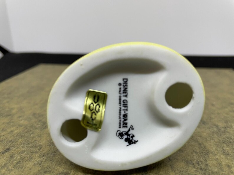 May include: A Disney Gift-Ware figurine base, featuring a yellow top and a white base with two holes. The base has the Disney Gift-Ware logo and a Mickey Mouse graphic. A gold sticker is attached.