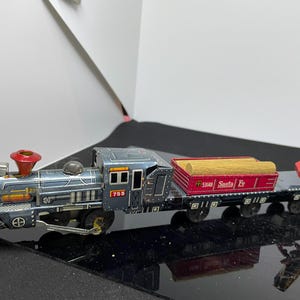 Könnte beinhalten: Ein Vintage-Zinn-Spielzeug-Zugsatz mit einer blauen Lokomotive, einem roten Güterwagen mit der Aufschrift "53440 Santa Fe" und einem gelben Kran. Der Zug hat einen roten Schornstein und schwarze Räder.