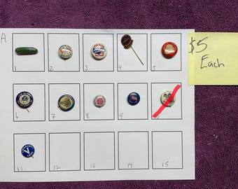 May include: A selection of vintage pins and buttons arranged on a white sheet. The pins showcase diverse designs, including text and graphics. A yellow sticky note indicates a price of £5 each.