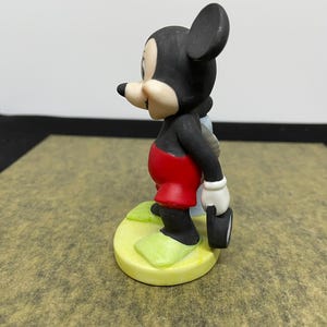 May include: A ceramic figurine of Mickey Mouse, a classic Disney character. Mickey is depicted in his iconic outfit: red shorts, black body, white gloves, and yellow shoes. He is holding a small, black object.