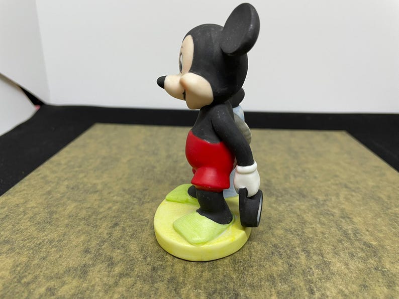 May include: A ceramic figurine of Mickey Mouse, a classic Disney character. Mickey is depicted in his iconic outfit: red shorts, black body, white gloves, and yellow shoes. He is holding a small, black object.
