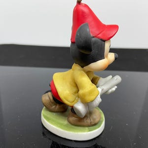 Puede incluir: Una figurita de porcelana de Mickey Mouse con un sombrero rojo y una chaqueta amarilla, sosteniendo un par de baquetas de bater&iacute;a plateadas.