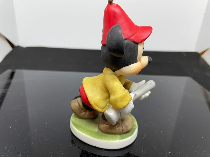Puede incluir: Una figurita de porcelana de Mickey Mouse con un sombrero rojo y una chaqueta amarilla, sosteniendo un par de baquetas de bater&iacute;a plateadas.