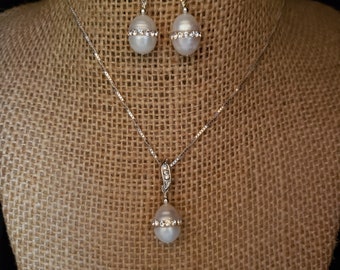 May include: A pearl necklace and earrings set with a silver chain and a single pearl pendant with a row of clear rhinestones around the pearl.