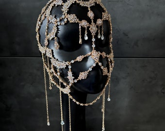 May include: A gold-toned headpiece with a delicate chain design and crystal embellishments. The headpiece features a face mask with a geometric pattern and dangling crystal drops.