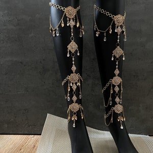 May include: A pair of gold leg chains with crystal accents. The chains are adorned with intricate designs and dangle from the thigh to the ankle.