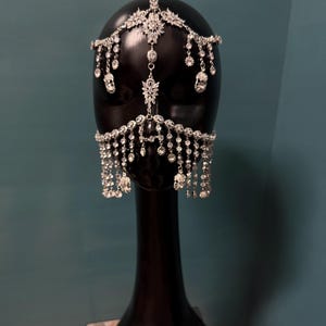 May include: A silver-toned headpiece with sparkling rhinestones and dangling accents. The ornate design features a central floral motif and cascading crystal strands, displayed on a black mannequin head. The headpiece is designed to be worn on the forehead and around the face.