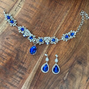 May include: A silver necklace with a blue crystal pendant and matching earrings. The necklace features a delicate floral design with smaller crystals.