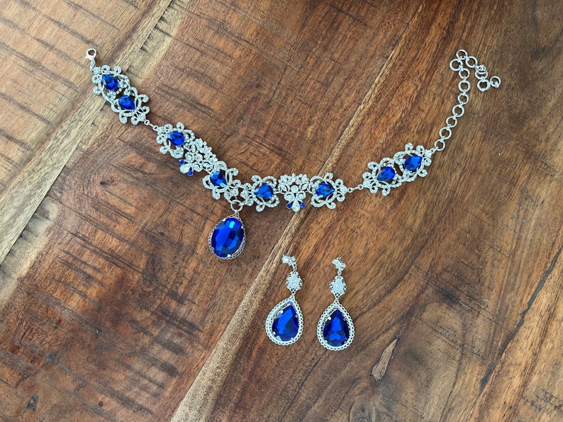 May include: A silver necklace with a blue crystal pendant and matching earrings. The necklace features a delicate floral design with smaller crystals.