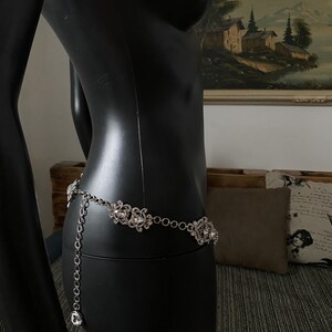 May include: A silver chain belt with large, clear rhinestone embellishments. The belt is on a black mannequin.