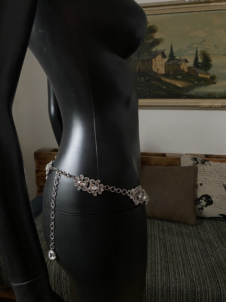 May include: A silver chain belt with large, clear rhinestone embellishments. The belt is on a black mannequin.