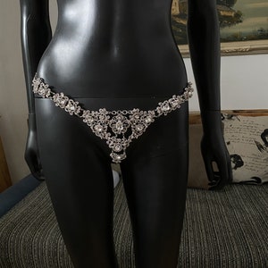 May include: A silver body chain with a delicate design featuring small, clear rhinestones. The chain is worn on a black mannequin.