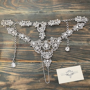 May include: A silver and crystal necklace with a delicate design. The necklace has a pendant that hangs down from the center. The necklace is on a wooden surface with a business card that says "GIVA MY JEWELRY".