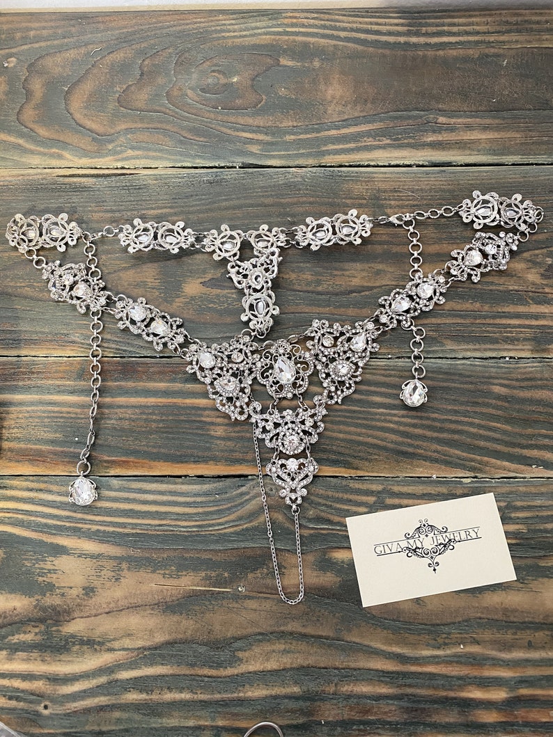 May include: A silver and crystal necklace with a delicate design. The necklace has a pendant that hangs down from the center. The necklace is on a wooden surface with a business card that says "GIVA MY JEWELRY".