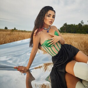May include: A woman wearing a green and white striped corset top with a black skirt and white boots. She is sitting on a reflective surface with a field of tall grass in the background.
