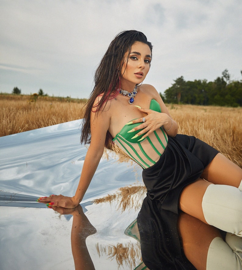 May include: A woman wearing a green and white striped corset top with a black skirt and white boots. She is sitting on a reflective surface with a field of tall grass in the background.