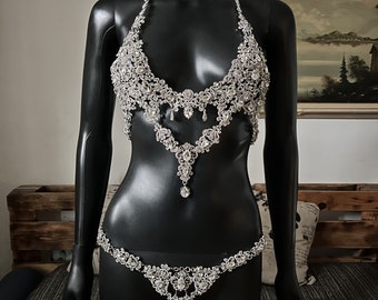 May include: A silver-toned body chain with a delicate, intricate design featuring small, clear rhinestones. The chain is worn on a black mannequin and includes a bra top and a matching bottom piece.