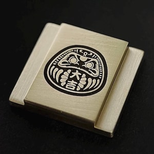 以下が含まれることがあります： 銀色の金属製の正方形で、黒と白の彫刻が施された日本のだるま人形の画像が描かれています。人形の額には「大」の漢字が書かれています。