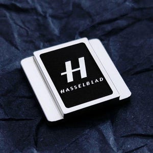 Może przedstawiać: Talia kart do gry z czarną kartą z białym logo "H" i napisem "HASSELBLAD" w kolorze białym. Karty są umieszczone na ciemnoniebieskim, teksturowanym tle.