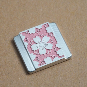 May include: A small, square, silver-coloured metal charm with a pink and white cherry blossom design.