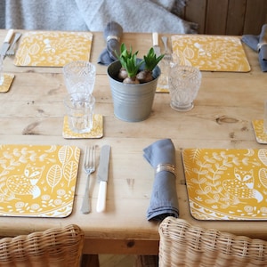 Puede incluir: Una mesa puesta con manteles individuales amarillos con un diseño de zorro blanco. Los manteles individuales están sobre una mesa de madera con una servilleta gris y un anillo de servilleta plateado. Hay una pequeña planta en maceta en el centro de la mesa.
