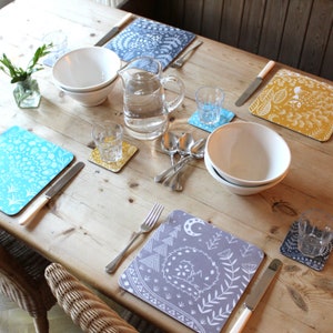 Puede incluir: Un juego de cuatro manteles individuales de patrón gris y blanco con un diseño de zorro, luna y estrellas. Los manteles individuales están sobre una mesa de madera con una jarra de agua, cuencos y vasos.