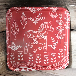 May include: A square, red serving tray with a white Dala horse design. The tray features a repeating pattern of stylized trees, flowers, and foliage. The tray is made of a glossy material and has rounded corners.