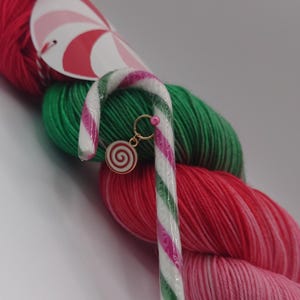 Op de afbeelding: Een stekenmarkeerder met een snoepstokthema, met een goudkleurige ring en een spiraalvormige bedel, tegen een achtergrond van rode, groene en roze wol. Het garen is in een gelaagd patroon gerangschikt, waardoor een feestelijke en kleurrijke weergave ontstaat.