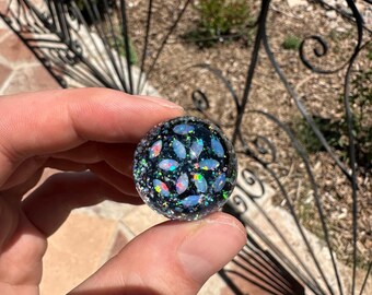 May include: A round, black marble with embedded iridescent opal-like pieces and glitter. The marble is held in a hand, with a wrought iron fence and stone patio in the background.