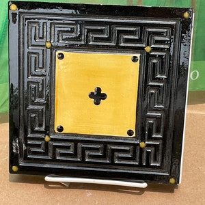 May include: A black and yellow ceramic tile with a Greek key border and a yellow center with a black cross. The tile has four gold dots on the border.