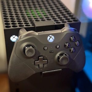 Peut inclure: Une console Xbox Series X noire avec une manette Xbox noire fixée à l'avant. La manette a un bouton Xbox blanc et la console a un voyant vert.